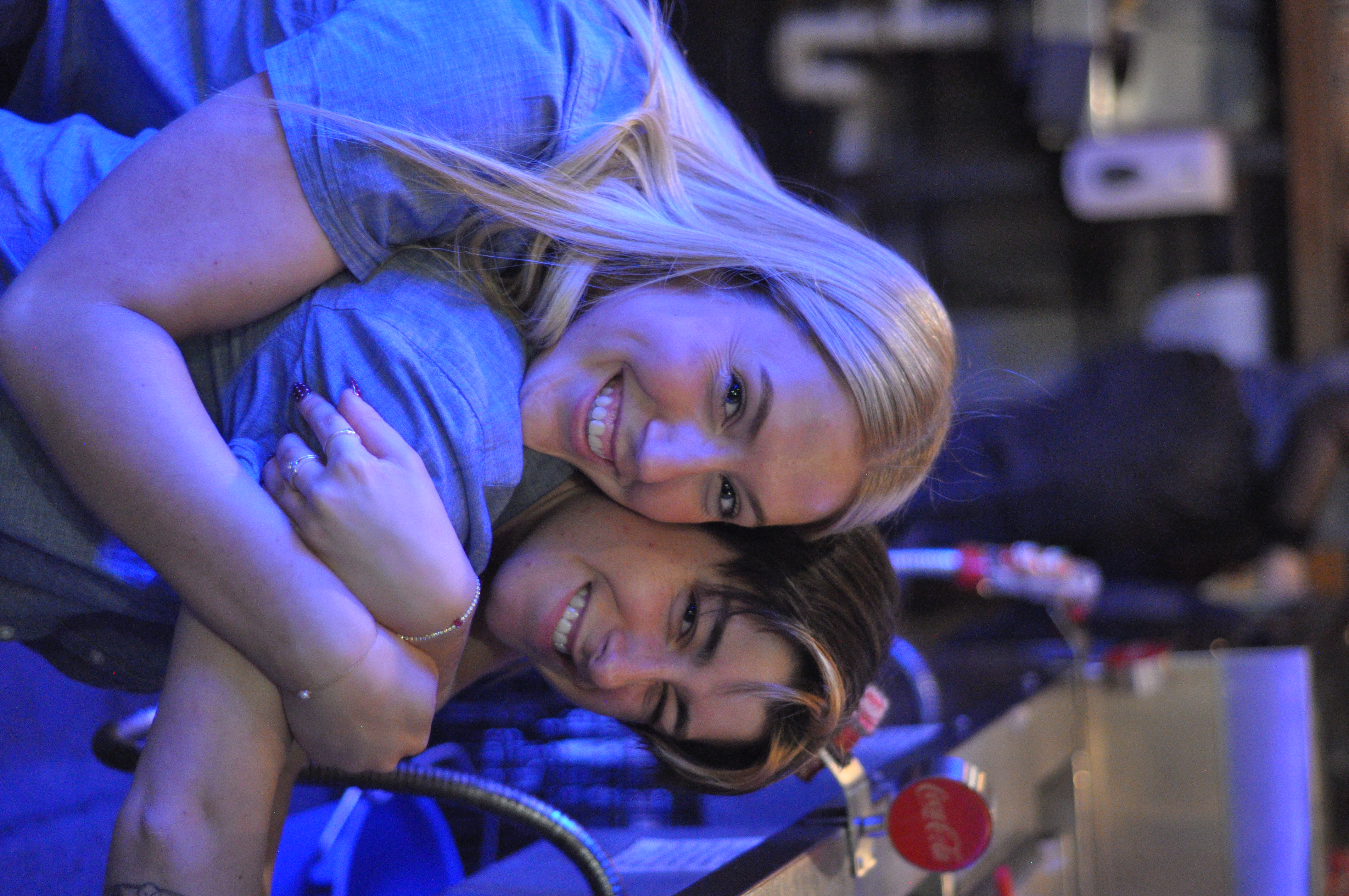 Two friends smiling together behind a bar under blue lighting